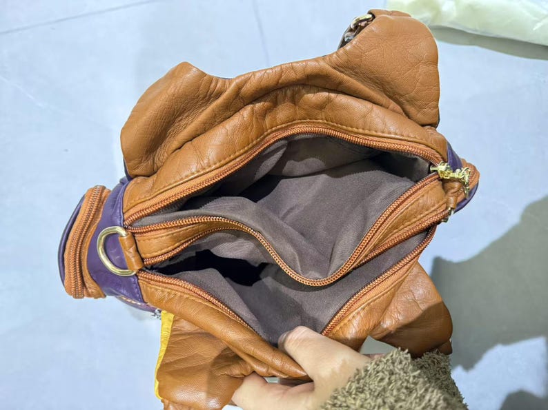 Puede incluir: Un bolso de mano de cuero marr&oacute;n con forro morado y varios compartimentos. El bolso tiene cierre de cremallera y una hebilla dorada.