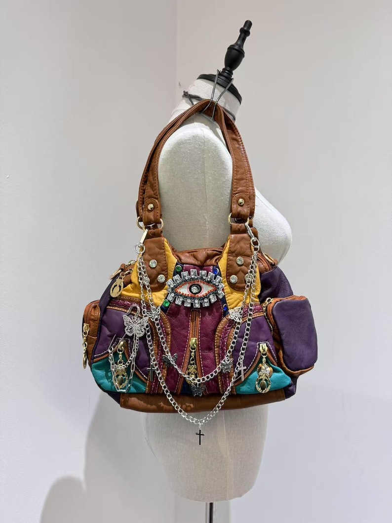 Puede incluir: Un bolso de mano de cuero multicolor con un gran dise&ntilde;o de ojo en la parte delantera. El bolso tiene varios bolsillos y est&aacute; adornado con cadenas y abalorios, incluyendo una mariposa, una cruz y un coraz&oacute;n.