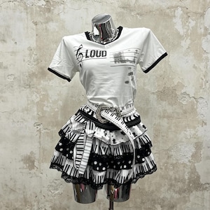Zweiteiliges Outfit im Y2K-Alternative-Scene-Kid-Gyaru-Stil: Kurzarm-Top mit Musiknoten-Print, dazu ein gepunkteter Minirock mit Rüschen und Klaviertastenmuster sowie ein gestufter Minirock mit Spitzenrüschen im Klaviertasten-Stil.