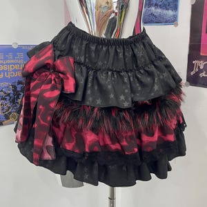 May include: A tiered skirt featuring black fabric with a subtle star pattern, alternating with layers of red and black animal print fabric, black faux fur trim, and black lace edging. A large matching red and black animal print bow is attached.