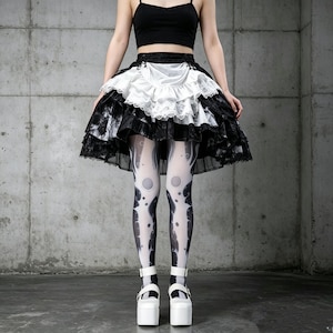 May include: A black sleeveless crop top paired with a layered black and white skirt featuring a ruffled white apron-style front with lace trim. The outfit includes white tights with a black abstract pattern and white platform shoes with ankle straps.