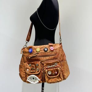 May include: A brown leather shoulder bag with multiple pockets and decorative embellishments, including buttons, pins, and a large eye design.
