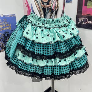 May include: A tiered skirt featuring alternating layers of mint green fabric with black star and cat prints and teal and black gingham check fabric. The skirt has an elastic waistband and is trimmed with black lace along each of the ruffled tiers.