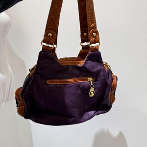 Puede incluir: Un bolso de mano de cuero morado con correas de cuero marr&oacute;n y un tirador de cremallera dorado. El bolso tiene un peque&ntilde;o bolsillo en la parte delantera.
