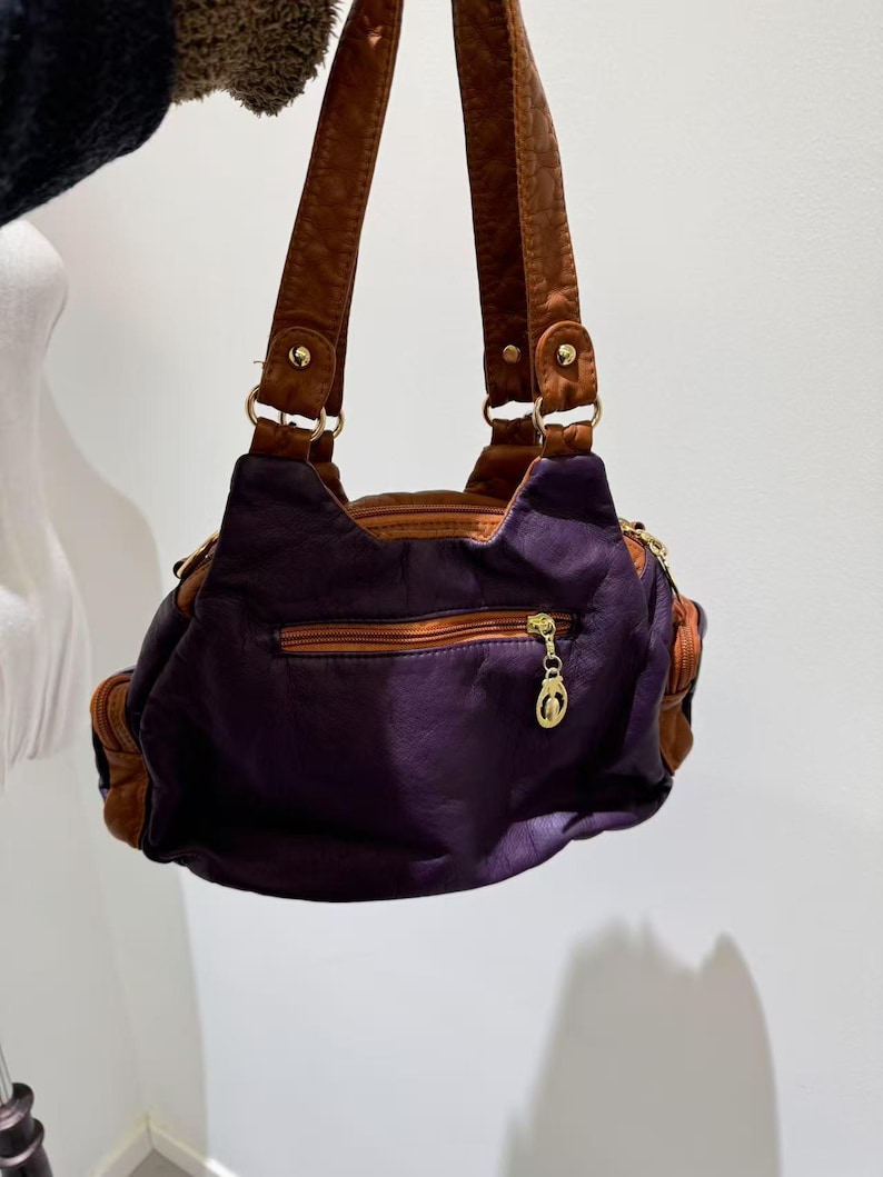 Puede incluir: Un bolso de mano de cuero morado con correas de cuero marr&oacute;n y un tirador de cremallera dorado. El bolso tiene un peque&ntilde;o bolsillo en la parte delantera.