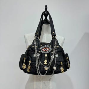 Puede incluir: Bolso de mano de cuero negro con un gran dise&ntilde;o de ojo adornado con pedrer&iacute;a y detalles dorados. El bolso tiene varios compartimentos con cremallera y est&aacute; adornado con cadenas colgantes y abalorios de mariposa.