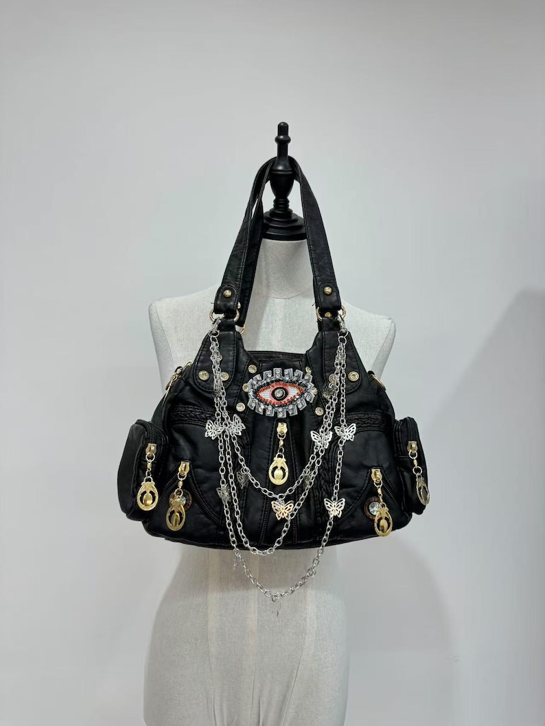 Puede incluir: Bolso de mano de cuero negro con un gran dise&ntilde;o de ojo adornado con pedrer&iacute;a y detalles dorados. El bolso tiene varios compartimentos con cremallera y est&aacute; adornado con cadenas colgantes y abalorios de mariposa.