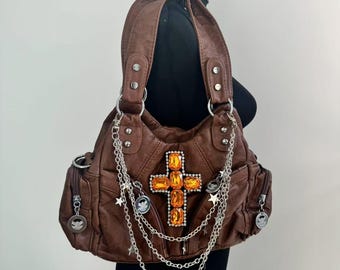 May include: A brown leather handbag with a large jeweled cross on the front. The bag has multiple silver chains and star charms hanging from it.