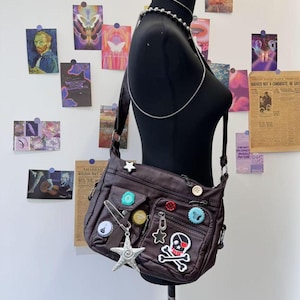 以下が含まれることがあります： カラフルなボタン、チャーム、ドクロとクロスボーンのパッチが付いた、茶色のフェイクレザーのショルダーバッグ。バッグにはジッパー付きポケットとフラップクロージャーがあります。