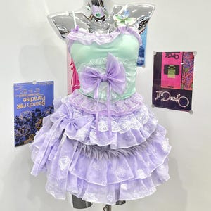 Peut inclure: Une robe sans manches avec un corsage vert menthe orné d'un grand nœud lavande au centre. La jupe se compose de multiples volants lavande superposés avec une bordure en dentelle blanche et un motif floral texturé, agrémentée d'une fourrure grise.