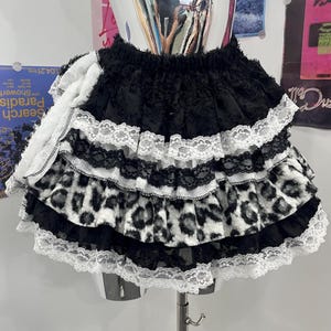 Può includere: Una gonna a balze a strati con alternanza di tessuto nero, bordi in pizzo bianco e una stampa leopardata in bianco e nero. La gonna presenta un elastico in vita e una silhouette voluminosa con molteplici balze orizzontali rifinite in pizzo.