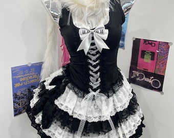 May include: A black sleeveless dress featuring a white faux fur collar, a front lace-up corset bodice with a piano key patterned bow, and a multi-layered skirt with alternating black and white lace ruffles and a long white faux fur tail.