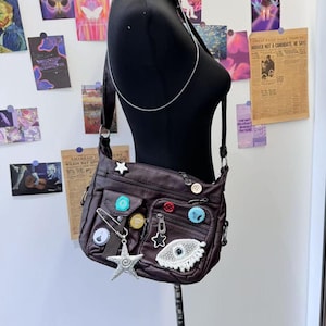 May include: A brown faux leather shoulder bag with multiple pockets and zipper closures. The bag is decorated with various colorful buttons, a silver star charm, and a beaded eye charm.