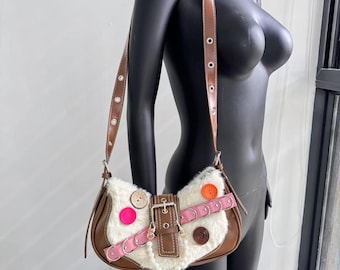 May include: A brown leather shoulder bag with a white faux fur flap. The flap has a buckle closure and is decorated with colorful buttons and pink ribbon.