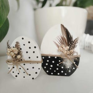 May include: Two white wooden eggs decorated with black polka dots and dried flowers. The eggs are sitting on a white surface.