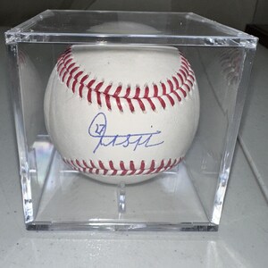 May include: A white baseball with red stitching and a blue signature, presented in a clear acrylic display case. The baseball is the focal point, with the signature and stitching clearly visible. The case is square and transparent.