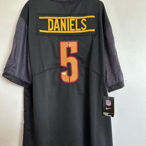 以下が含まれることがあります： 黒色のフットボールジャージ。名前「DANIELS」は黄色の文字で、番号「5」は赤と黄色で表示されています。ジャージはグレーのメッシュスリーブとNFLタグが付いています。