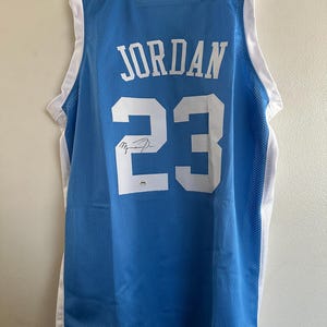 以下が含まれることがあります： 白のトリムが付いた水色のバスケットボールジャージ。名前「JORDAN」と番号「23」は白です。印刷された署名が含まれています。ジャージは白いハンガーに吊るされています。