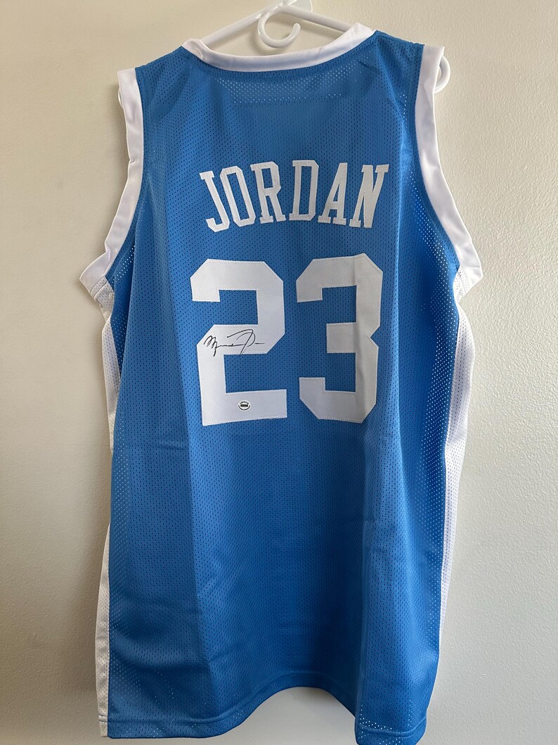 以下が含まれることがあります： 白のトリムが付いた水色のバスケットボールジャージ。名前「JORDAN」と番号「23」は白です。印刷された署名が含まれています。ジャージは白いハンガーに吊るされています。
