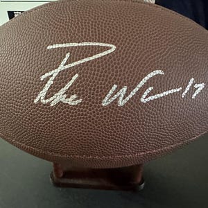 Puede incluir: Un balón de fútbol americano de cuero marrón con letras blancas que dicen "The Wc - 17". El balón está sobre un soporte marrón. El balón tiene una superficie texturizada.