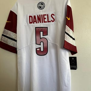 May include: A white Washington Commanders football jersey featuring the name DANIELS in red letters above the number 5 in red with a white and black outline. The sleeves are burgundy with white and black accents and a yellow Nike swoosh logo.