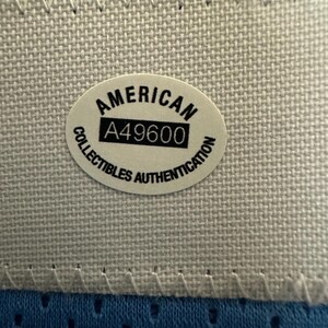 以下が含まれることがあります： 白いステッチの縁取りが付いた青い生地のクローズアップ。 「AMERICAN」と「A49600」の文字が入った楕円形のステッカーが生地の中央に配置されています。 ステッカーには「COLLECTIBLES AUTHENTICATION」の文字も含まれています。