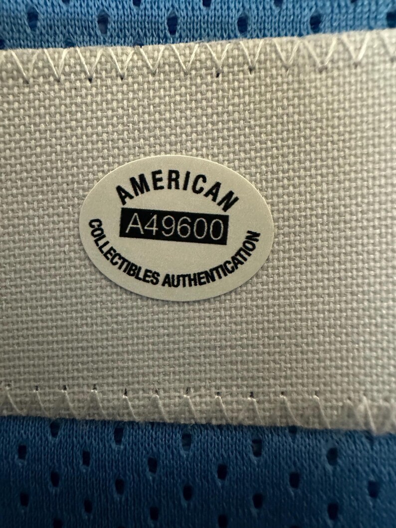 以下が含まれることがあります： 白いステッチの縁取りが付いた青い生地のクローズアップ。 「AMERICAN」と「A49600」の文字が入った楕円形のステッカーが生地の中央に配置されています。 ステッカーには「COLLECTIBLES AUTHENTICATION」の文字も含まれています。