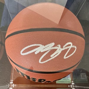 May include: A brown leather basketball with black rubber seams and a textured surface. The ball features a prominent white signature written in cursive across the center and the black Wilson brand logo printed near the bottom edge.