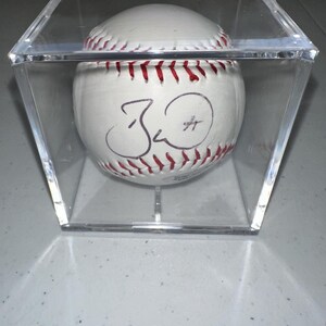 May include: A white baseball with red stitching and a black signature, presented in a clear acrylic case. The baseball sits on a clear stand, offering a protective display for the item.
