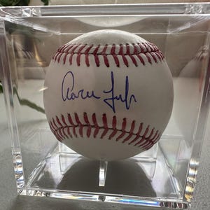 May include: A white baseball with red stitching and a blue signature, presented in a clear acrylic display case. The baseball is centrally positioned, highlighting the signature. The case is square and transparent.