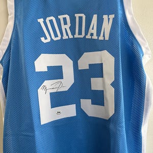 以下が含まれることがあります： 「JORDAN」の文字と数字の「23」が白でプリントされた水色のバスケットボールジャージ。ジャージには白いトリムと番号の上にサインが入っています。ジャージは通気性の良いメッシュ素材で作られています。