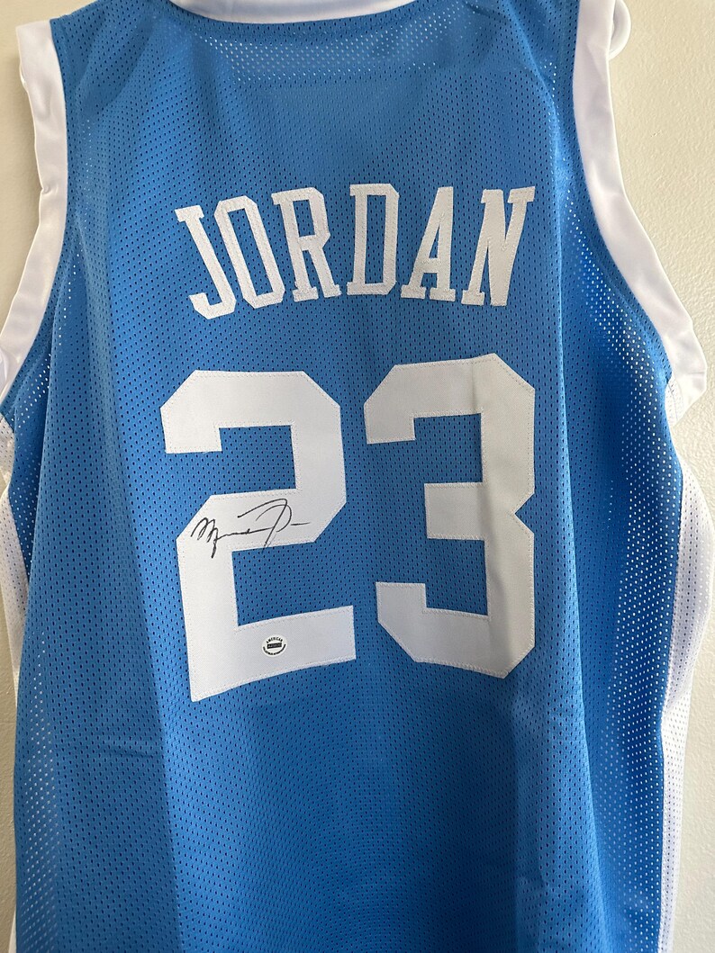 以下が含まれることがあります： 「JORDAN」の文字と数字の「23」が白でプリントされた水色のバスケットボールジャージ。ジャージには白いトリムと番号の上にサインが入っています。ジャージは通気性の良いメッシュ素材で作られています。