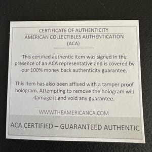 以下が含まれることがあります： 白地に黒文字の証明書。証明書はAmerican Collectibles Authentication (ACA)からのものです。テキストには、真正性の保証とウェブサイトのアドレスが含まれています。証明書は暗い表面にあります。