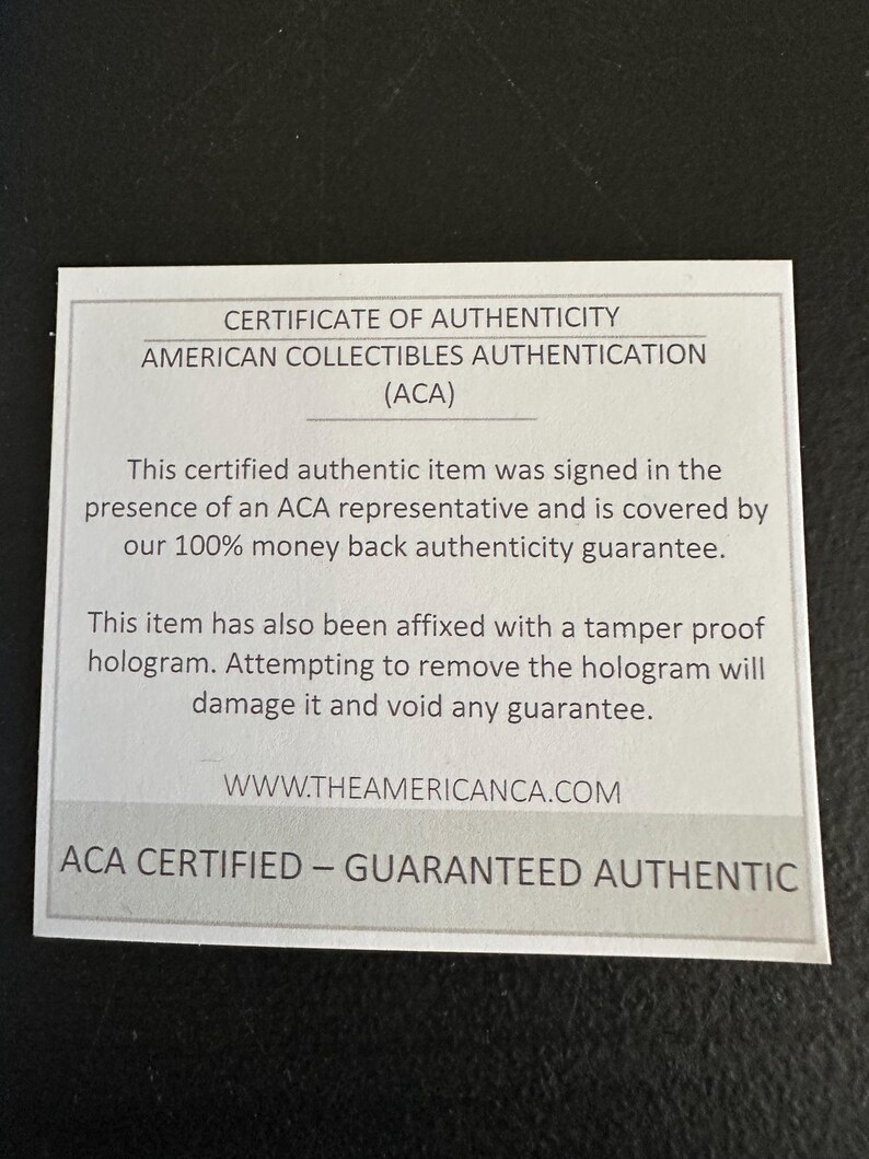 以下が含まれることがあります： 白地に黒文字の証明書。証明書はAmerican Collectibles Authentication (ACA)からのものです。テキストには、真正性の保証とウェブサイトのアドレスが含まれています。証明書は暗い表面にあります。