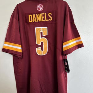 May include: Maroon football jersey with the name "DANIELS" and the number "5" in gold. The jersey has gold and white stripes on the sleeves and a Nike tag.