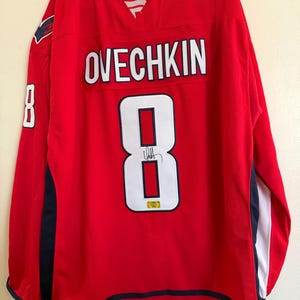 May include: A red hockey jersey featuring the name OVECHKIN and the number 8 in white with navy blue outlines on the back. The jersey includes a navy blue collar, navy blue side panels, and a white stripe on the sleeves. A signature is on the number 8.