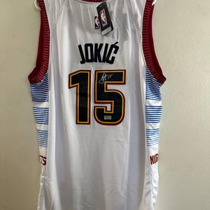 May include: A white Denver Nuggets basketball jersey featuring the name JOKIĆ and the number 15 in dark blue with yellow and red outlines. The jersey has maroon trim on the collar and armholes, with blue and white horizontal stripes on the sides.