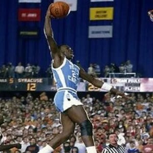 以下が含まれることがあります： 青と白のユニフォームを着たバスケットボール選手が、ボールをダンクするためにジャンプしています。選手は空中にいて、ボールを頭上に高く掲げています。大勢の人が試合を見ています。