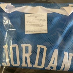 以下が含まれることがあります： 大きな白い文字で「JORDAN」と書かれた、水色のメッシュジャージ。ジャージは白い襟付きで、透明なビニール袋で密封されています。白と黒の真正性証明書が見えます。
