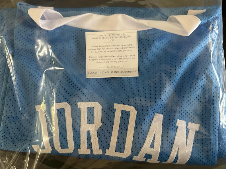 以下が含まれることがあります： 大きな白い文字で「JORDAN」と書かれた、水色のメッシュジャージ。ジャージは白い襟付きで、透明なビニール袋で密封されています。白と黒の真正性証明書が見えます。