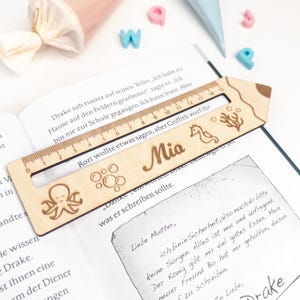 Personalisierte Lesehilfe aus Holz mit Lineal liegt auf einem geöffneten Kinderbuch. Das Leselineal ist mit dem Namen „Mia“ sowie Meer-Motiven wie Oktopus, Seepferdchen, Blasen und Koralle graviert – ideal als Geschenk zur Einschulung und als Inhalt für die Schultüte.
