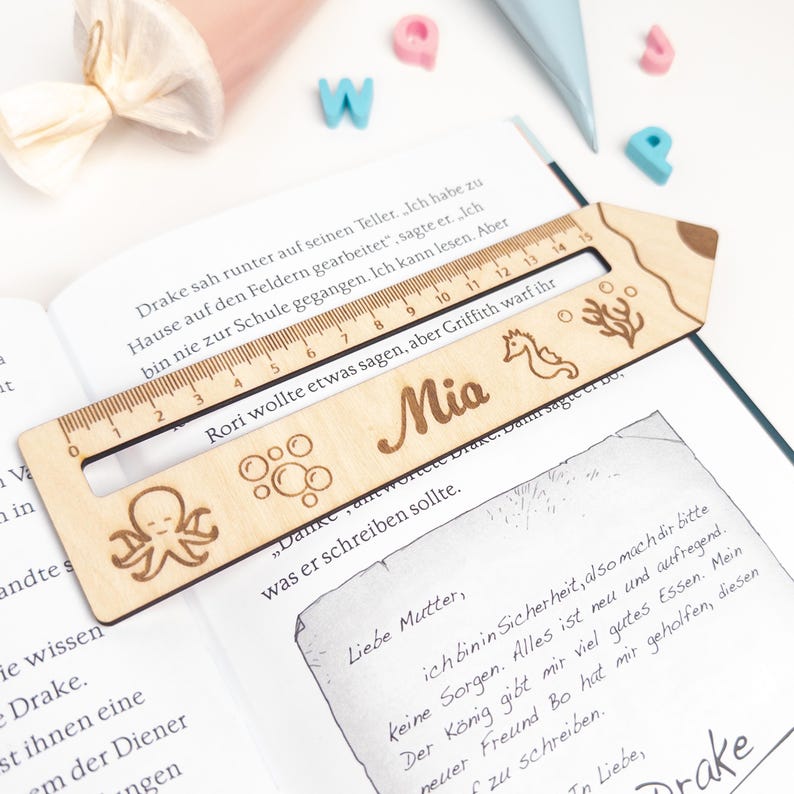 Personalisierte Lesehilfe aus Holz mit Lineal liegt auf einem geöffneten Kinderbuch. Das Leselineal ist mit dem Namen „Mia“ sowie Meer-Motiven wie Oktopus, Seepferdchen, Blasen und Koralle graviert – ideal als Geschenk zur Einschulung und als Inhalt für die Schultüte.