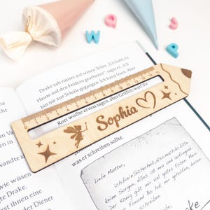 Personalisierte Lesehilfe aus Holz mit Lineal liegt auf einem geöffneten Kinderbuch. Das Leselineal ist mit dem Namen „Sophia“, einer kleinen Fee, Herz und Sternen graviert – ideal als Geschenk zur Einschulung oder als Inhalt für die Schultüte.