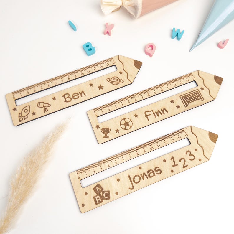 Drei personalisierte Lesehilfen aus Holz mit Lineal liegen auf hellem Hintergrund. Die Leselineale sind mit den Namen „Ben“, „Finn“ und „Jonas“ sowie Weltraum-, Fußball- und ABC-Motiven graviert – ideal als Geschenk zur Einschulung oder als Inhalt für die Schultüte.
