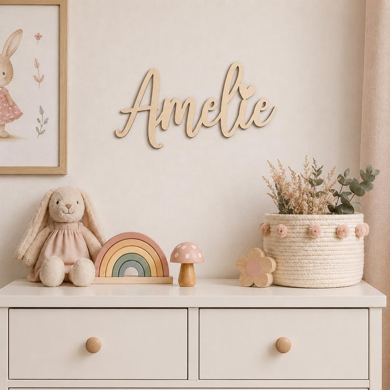 May include: A nursery scene with a wooden name sign that reads "Amelie" in cursive script. A plush bunny, a wooden rainbow, and a mushroom are on a white dresser. A framed bunny print and a woven basket with flowers complete the decor.