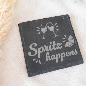 Ein quadratischer Untersetzer aus schwarzem Schiefer mit graviertem Spruch:  Spritz happens mit Gläsern und Zitrone. Der Untersetzer liegt dekorativ auf einem hellen Stofftuch.