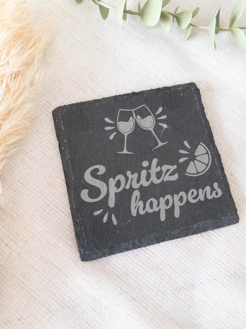 Ein quadratischer Untersetzer aus schwarzem Schiefer mit graviertem Spruch:  Spritz happens mit Gläsern und Zitrone. Der Untersetzer liegt dekorativ auf einem hellen Stofftuch.