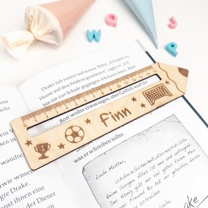 Personalisierte Lesehilfe aus Holz mit Lineal liegt auf einem geöffneten Kinderbuch. Das Leselineal ist mit dem Namen „Finn“, Fußball, Pokal, Sternen und Tor graviert – eine schöne Geschenkidee zur Einschulung und perfekt als Inhalt für die Schultüte.