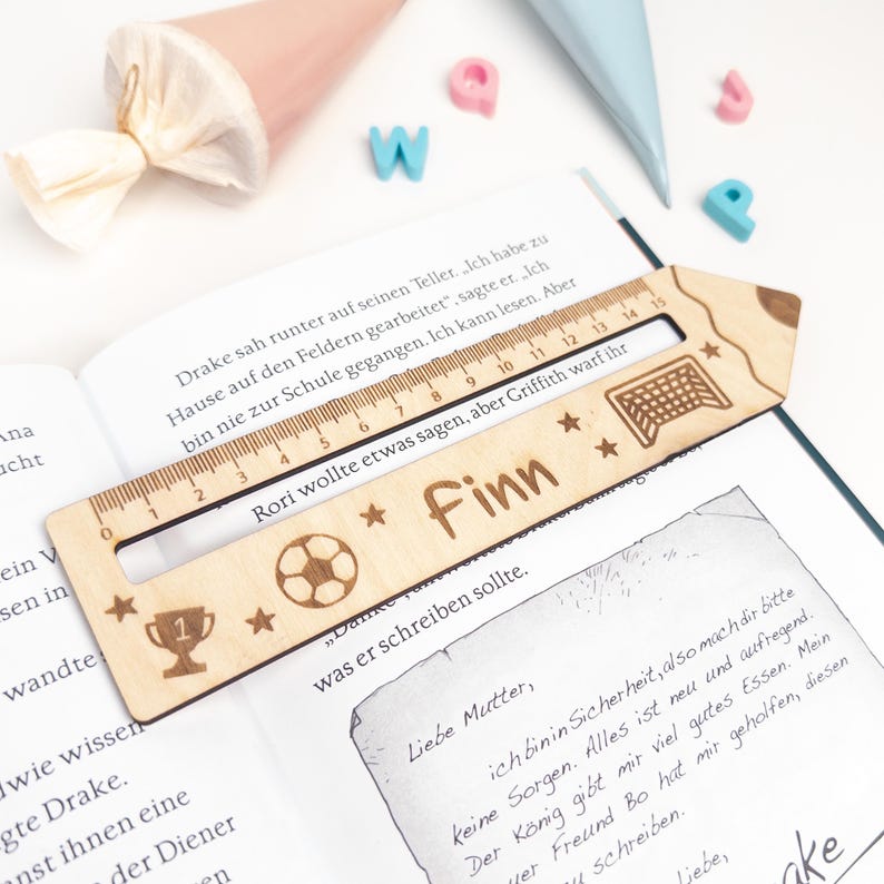 Personalisierte Lesehilfe aus Holz mit Lineal liegt auf einem geöffneten Kinderbuch. Das Leselineal ist mit dem Namen „Finn“, Fußball, Pokal, Sternen und Tor graviert – eine schöne Geschenkidee zur Einschulung und perfekt als Inhalt für die Schultüte.