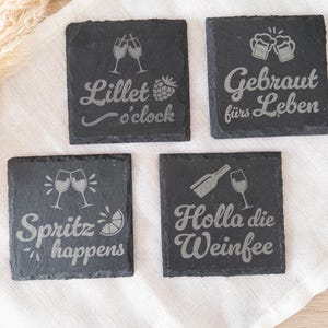 Vier quadratische Untersetzer aus schwarzem Schiefer mit gravierten Sprüchen: Lillet o’clock mit Gläsern und Himbeere, Gebraut fürs Leben mit Bierkrügen, Spritz happens mit Gläsern und Zitrone sowie Holla die Weinfee mit Flasche und Weinglas. Die Untersetzer liegen dekorativ auf einem hellen Stofftuch.